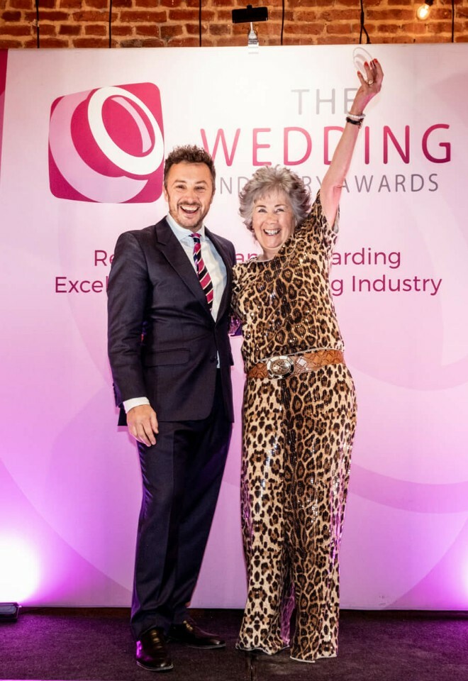 Suffolk Celebrant dressed in a leopard print trousers suit holds her trophy high. Next to her stands the male owner of the awards in a dark suit. He is smiling too