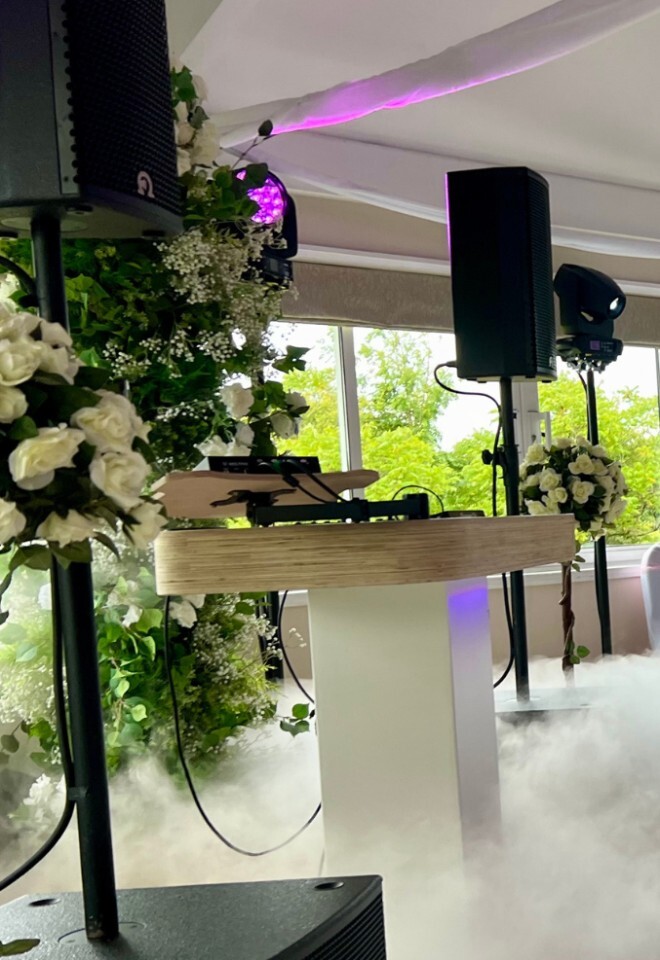 A bride and groom sharing their first dance on a dance floor covered in low-lying fog, with a three-tier wedding cake and a floral wall featuring a glowing "Mr & Mrs" neon sign in the background.