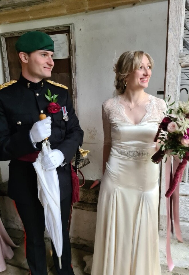 Bride wearing soft natural bridal makeup with satin wedding dress, standing beside groom in military uniform holding bouquet