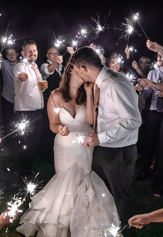 “Bride and groom kissing during a sparkler exit at a night wedding at Moreton Village Hall in Dorset by Katy Brothers Wedding Photography”