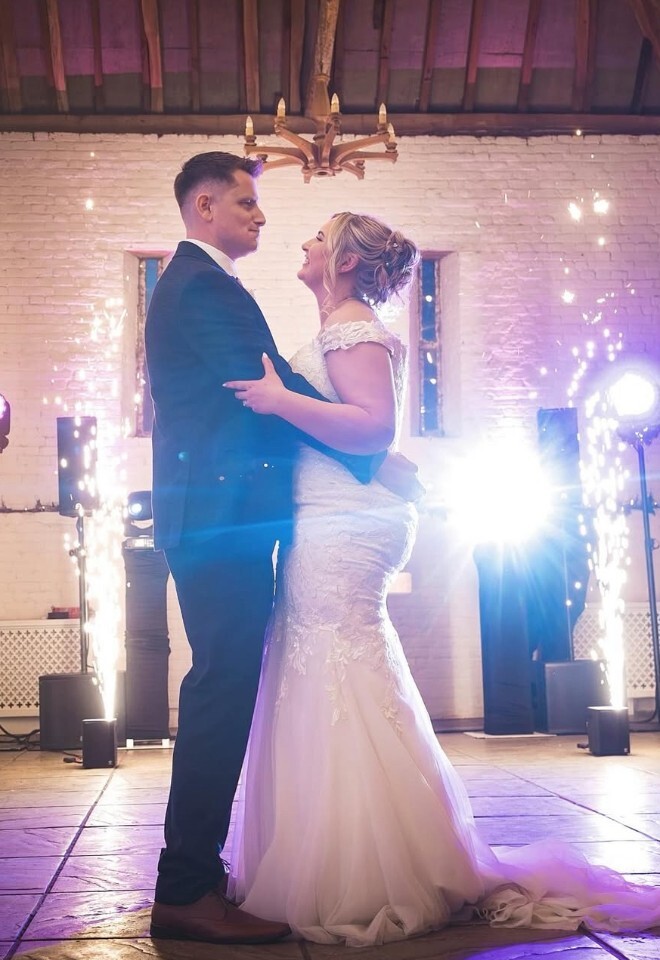 A newlywed couple embracing during their first dance in a rustic hall, framed by indoor sparkler fountains and bright stage lights under a wooden beam ceiling.