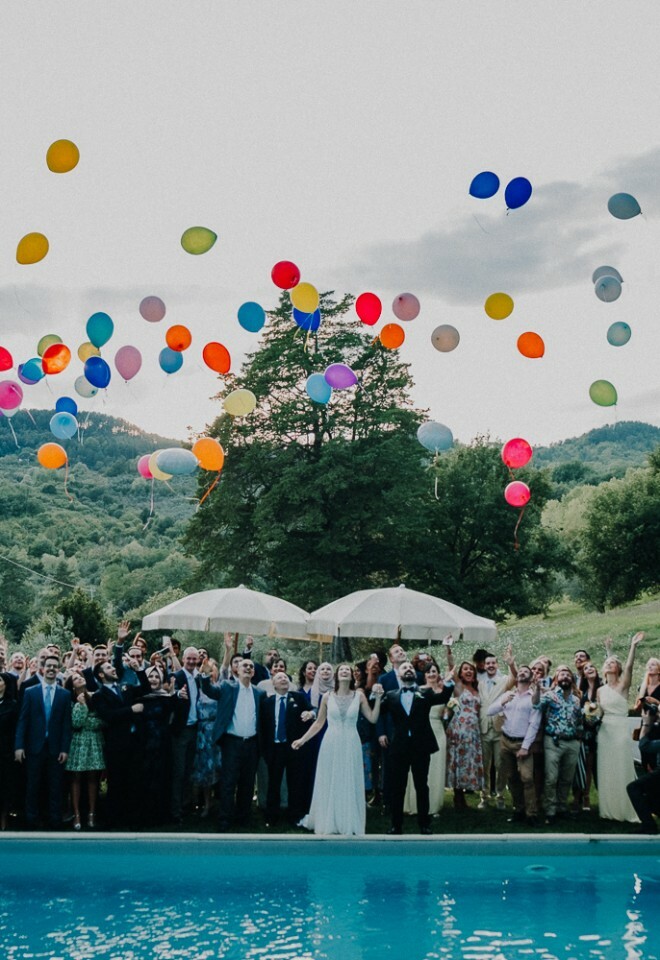 Wedding guests release colorful balloons into the sky during a joyful outdoor celebration by the pool, surrounded by lush greenery – documentary-style wedding photography