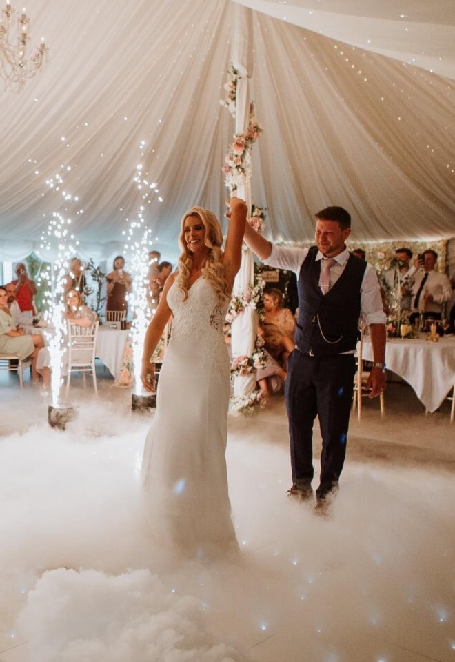 A newlywed couple smiling and holding hands during their first dance on a fog-covered dance floor, surrounded by sparkling indoor fountains and wedding guests inside a decorated marquee tent.