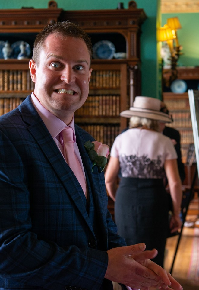 Terrified groom grimaces at the camera as he waits at the top of the aisle for his bride to enter in the Library at Prestwold Hall, Loughborough