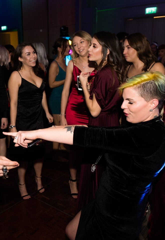 A lively crowd dancing at a wedding, featuring a bald man in a sparkly tuxedo jacket dancing across from a woman with short, yellow-tipped hair who is pointing forward.