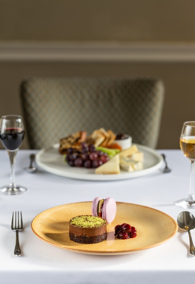 Elegant dessert and cheese course at Washingborough Hall Hotel, featuring chocolate pistachio cake, macaron, cherries, and wine on a white tablecloth.