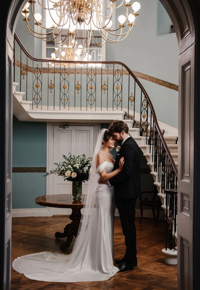 Bride and groom embracing beneath the sweeping staircase at Henlade House wedding venue in Somerset, showcasing classic interiors.