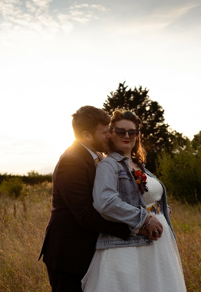 Bride and groom embracing at sunset in the middle of an overgrown field, bride is wearing a denim jacket and heart sunglasses