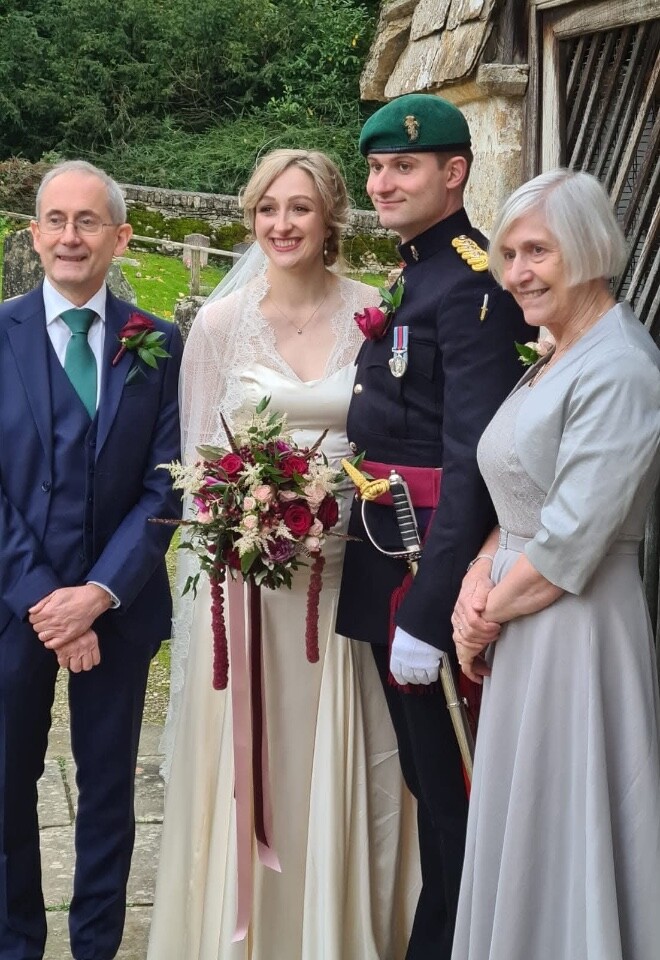Bride with soft natural bridal makeup standing with groom in military uniform and family, holding bouquet.