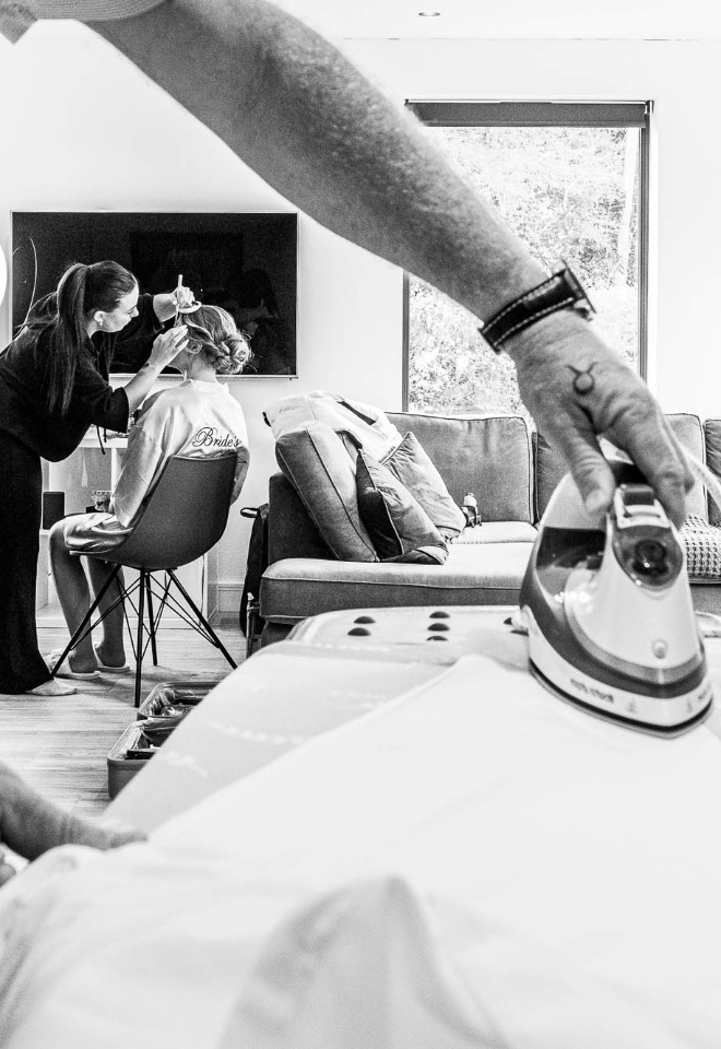 Father of bride ironing his shirt in the foreground whilst the bride is having her makeup applied in the background