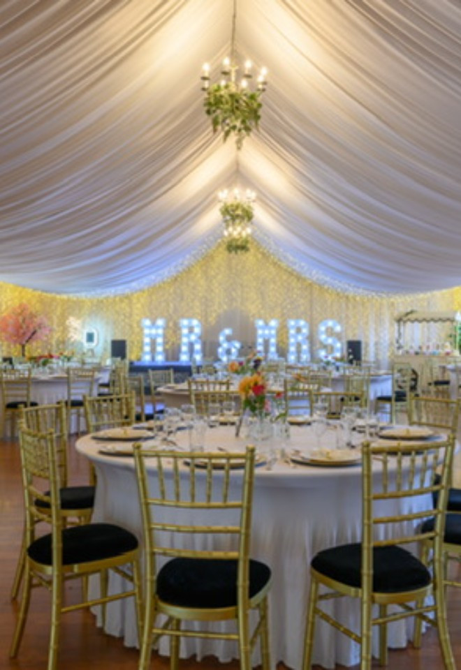 Wedding reception at Duvale Priory in Tiverton, Devon, with elegant round tables, gold chairs, draped ceilings, chandeliers and illuminated MR & MRS letters.