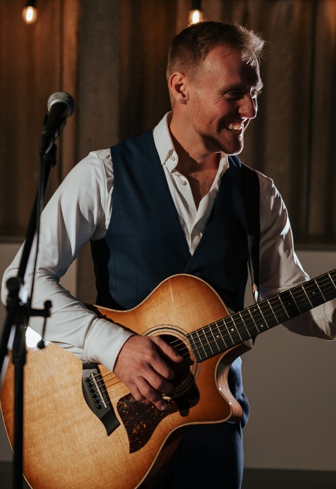 Matt Rayner, an Essex based guitarist and singer is laughing while playing guitar in a navy waistcoat