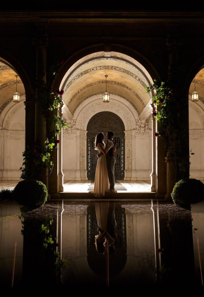 Bride and groom silhouetted at night beneath the lit stone arches of the Loggia at North Mymms Park, reflected in the water feature in front.