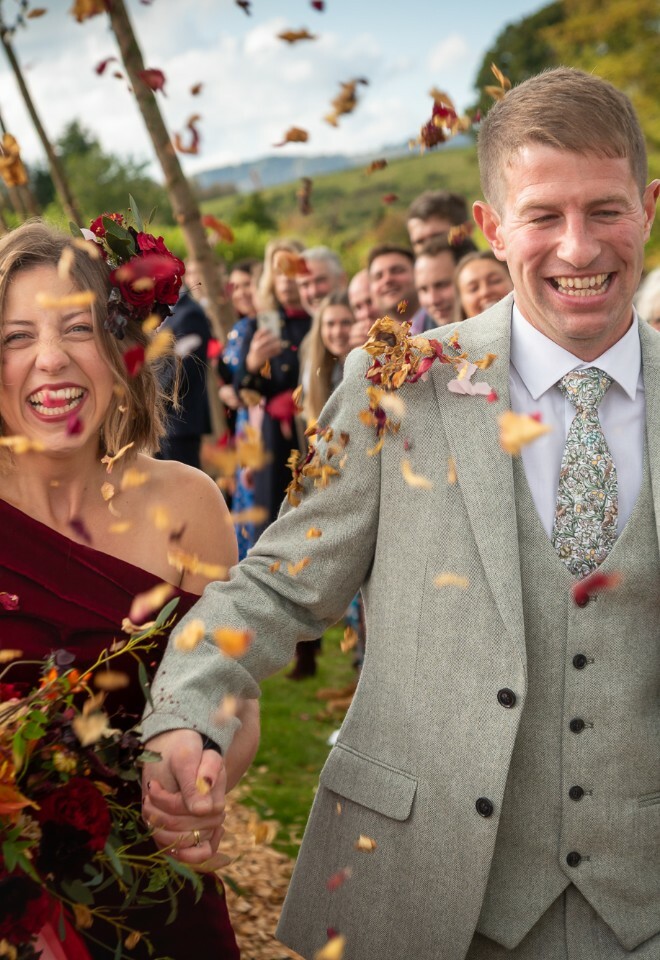 Newlyweds walk through shower of confetti with joyful wedding guests in garden setting.
