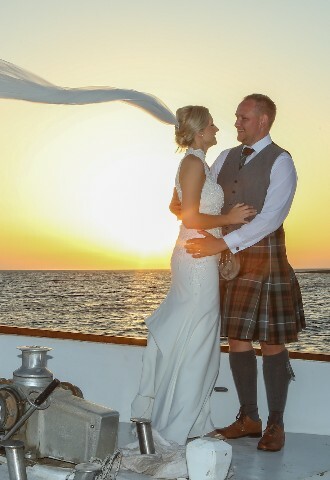 Bride and groom, in a kilt, pose at sunset at the front of a yacht in cyprus