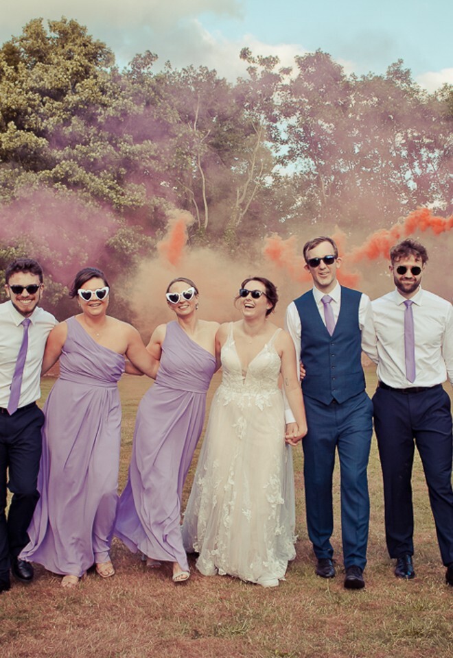 Bridal party walking towards camera wearing sunglasses and with coloured smoke in the background