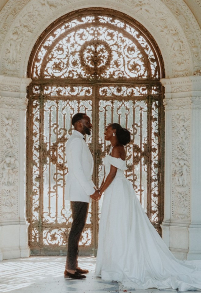African bride and groom holding hands inside the Loggia at North Mymms Park, positioned in front of intricate wrought-iron gates, flanked by white carved stone walls and potted palm trees.