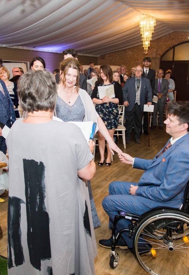 Wedding ceremony inside a decorated reception venue with greenery archways, guests gathered around, and a groom in a wheelchair holding hands with their partner during the vows.