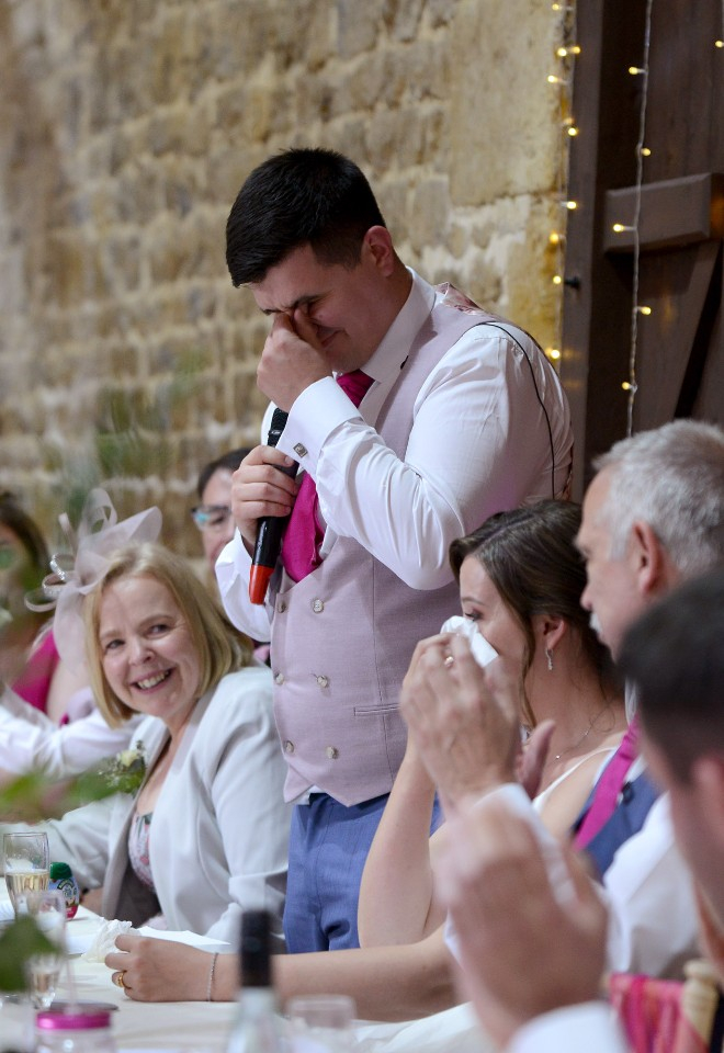 Groom covering his face during the wedding speeches as bride seated next to him uses her napkin to dab tears of laughter from her eyes and mother-in-law looks on smiling