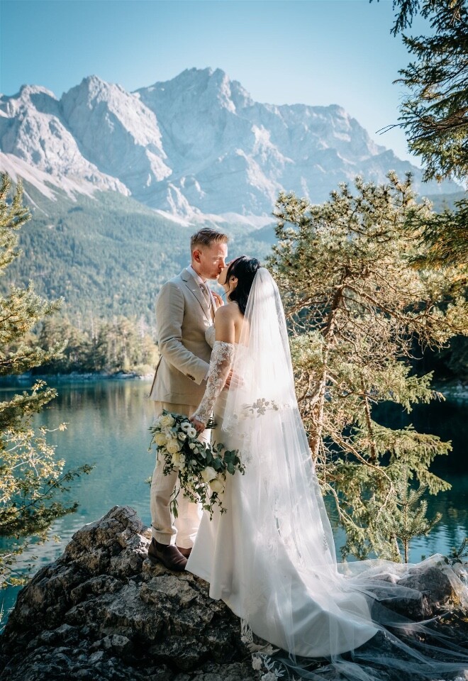 Lake Eibsee a glacial lake in Germanys Alps is where this bride and groom made their wedding vows. They are standing facing each other and smiling. The lake is behind them with a mountain backdrop 