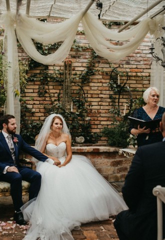 wedding couple sitting in a flower nursery with greenery all around them while the Celebrant is reading the ceremony script.