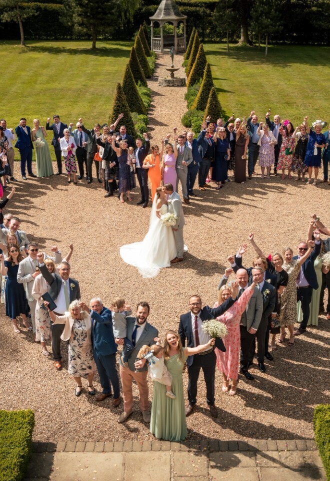 Wedding guests form a heart shape around the bride and groom kissing in the gardens of Washingborough Hall Hotel, celebrating under bright afternoon sunshine.
