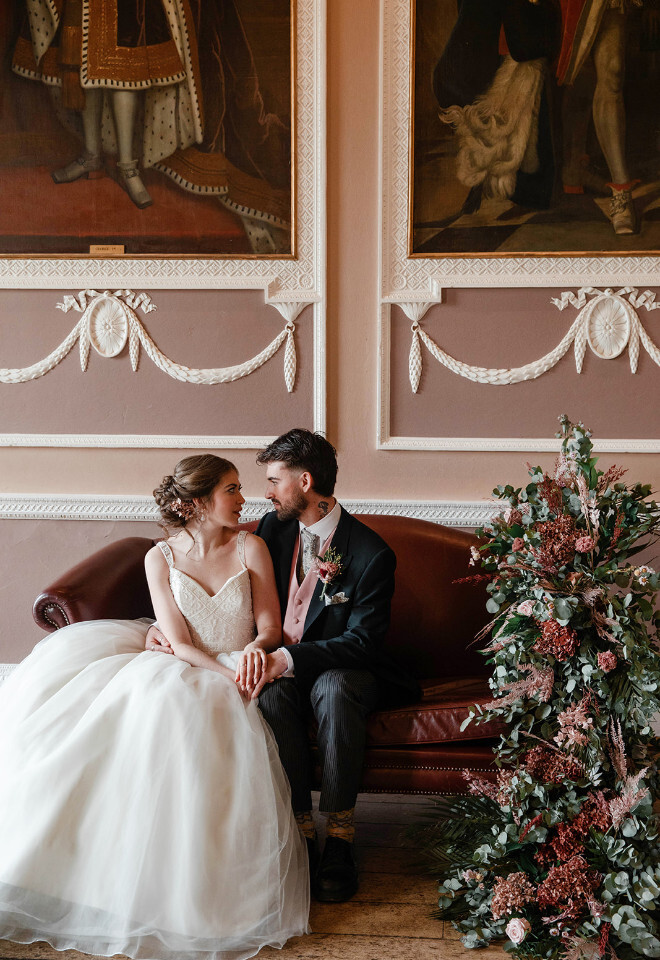 “Bride and groom sitting on sofa inside Milton Abbey Dorset with ornate wall details and floral arrangement”
