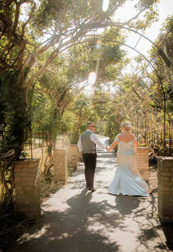 Bride and groom walking hand in hand under a sunlit garden archway