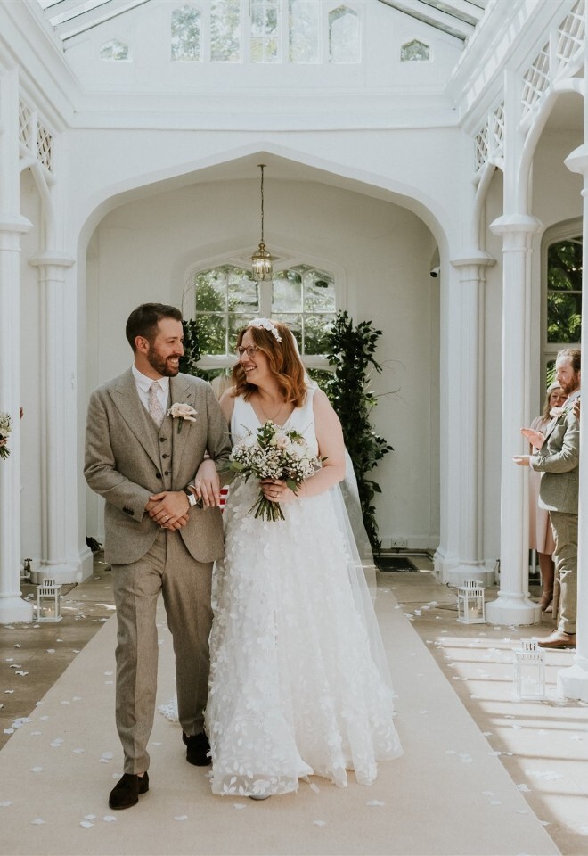 Bride and groom walking down orangery aisle with dusky pink and white wedding flowers at stately home venue by FLOWERS BY ANYA