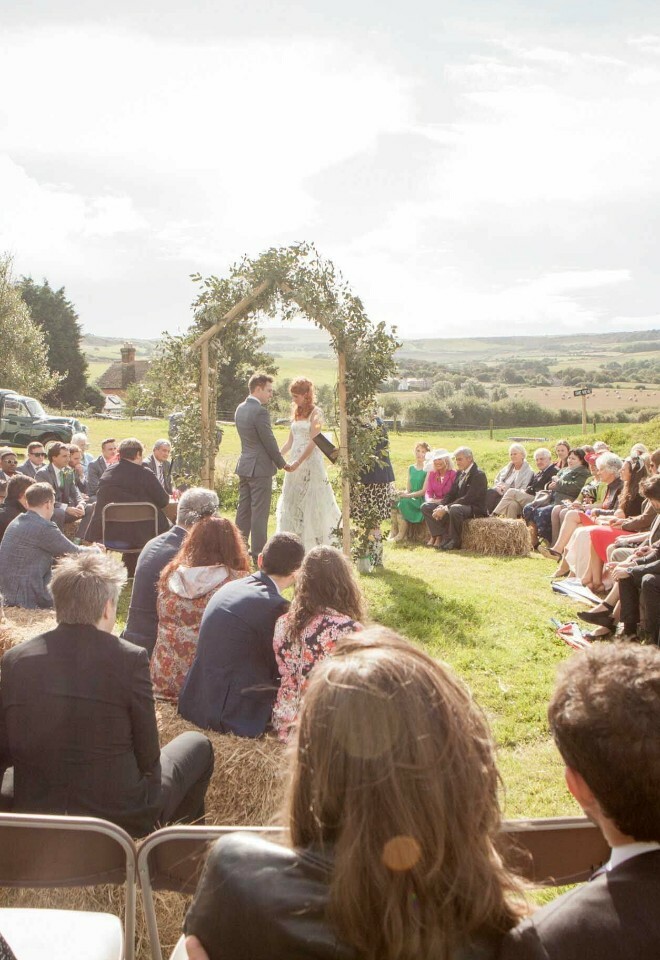 Rustic outdoor wedding ceremony with floral arch and guests on hay bales