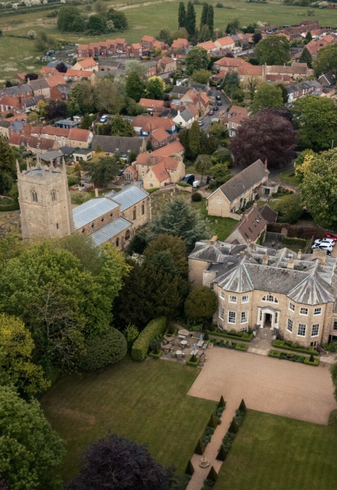 Aerial view of Washingborough Hall Hotel and St John the Evangelist Church surrounded by trees, gardens, and village houses in Lincolnshire countryside.