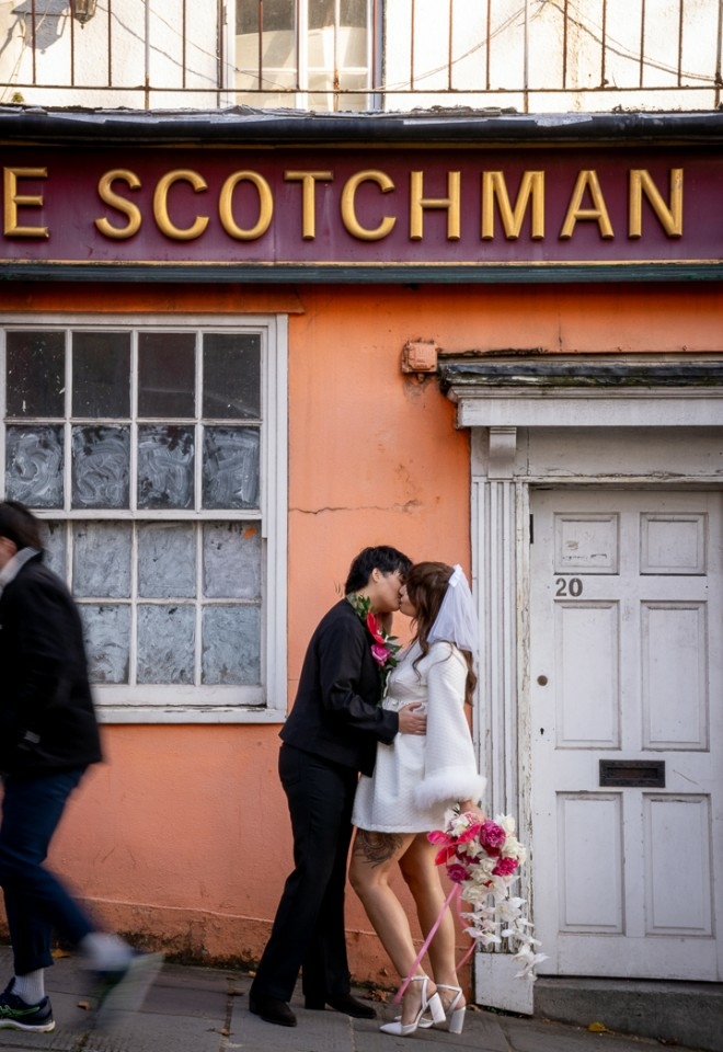 Brides kissing on the city streets, first bride wearing a suit a boots, second bride with a short vintage white dress