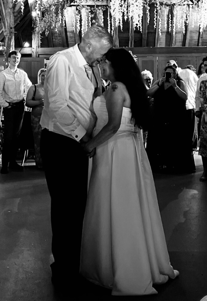 A black and white photo of a bride and groom touching foreheads during their first dance in a rustic barn venue, surrounded by wedding guests watching them.