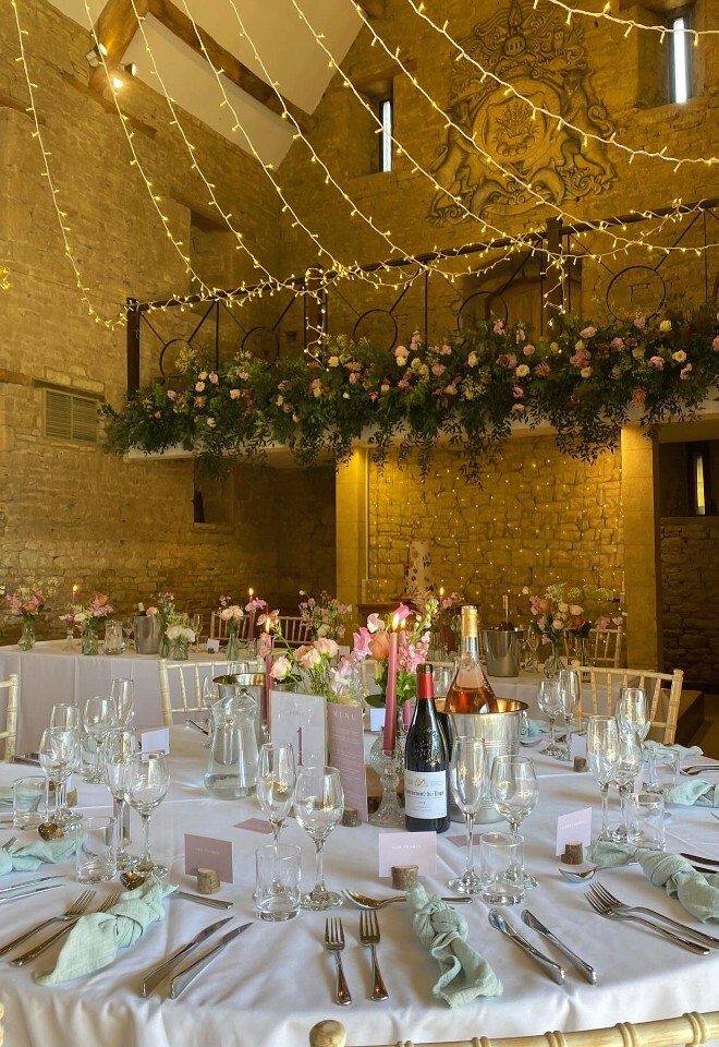 Wedding reception setup inside Great Tythe Barn with long tables dressed in white linens, glassware and soft green napkins, floral centrepieces, wine bottles, and warm fairy lights draped across