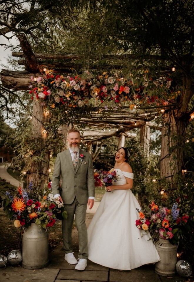 bride and groom stand beneath a rustic wooden pergola decorated with hanging fairy lights and a lush, colorful floral arrangement large milk churns line the stone pathway around them at Bennetts farm