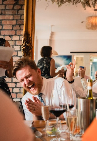 A groom looks shocked while his new husband laughs at a joke in the wedding speeches