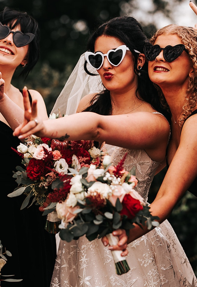 Bride posing with her bridesmaids in heart-shaped sunglasses, holding colourful wedding bouquets at The Cock O’Barton.