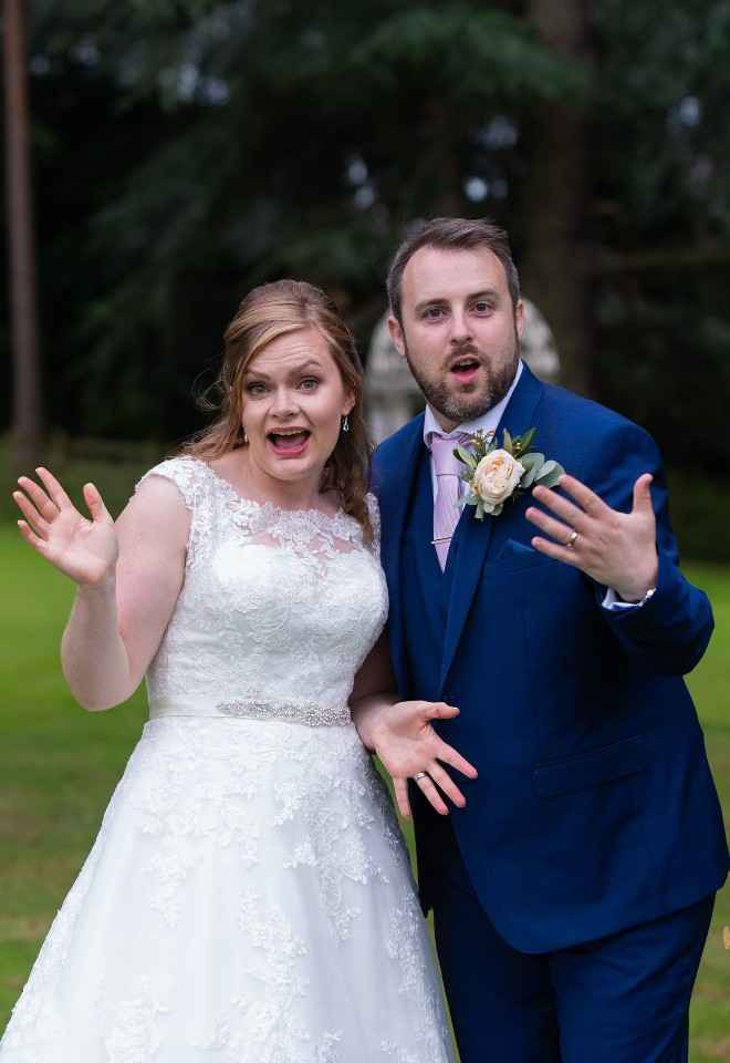 Newly married bride and groom show off their wedding rings whilst doing jazz hands and looking delighted to camera in the gardens of Sutton Bonington Hall