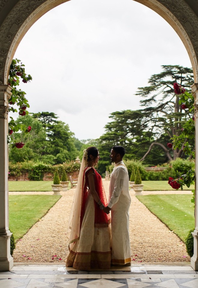 Bride and groom in traditional South Asian wedding attire holding hands framed by the stone archway of the Loggia at North Mymms Park, looking out over the landscaped gardens
