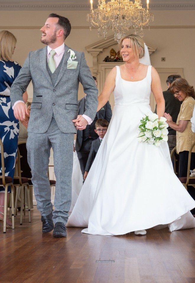 A wedding reception moment showing a couple walking down the aisle between rows of chairs in an elegant venue with chandelier.