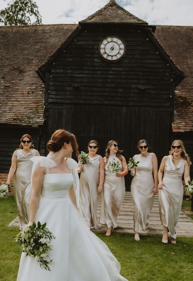 Bridesmaids in champagne satin walk with the bride outside a barn wedding venue in Oxfordshire, photographed by V & H Photography.