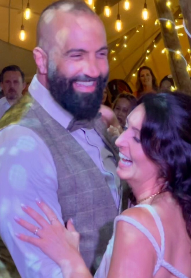 A happy bride and groom laughing together during their first dance under warm string lights inside a teepee marquee tent, surrounded by smiling wedding guests.