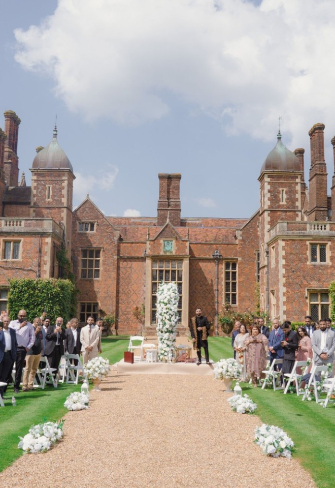 Outdoor Nikkah ceremony setup on the lawn at North Mymms Park, featuring guest seating flanking a central gravel walkway with white floral arrangements, set against the backdrop of the historic red-br