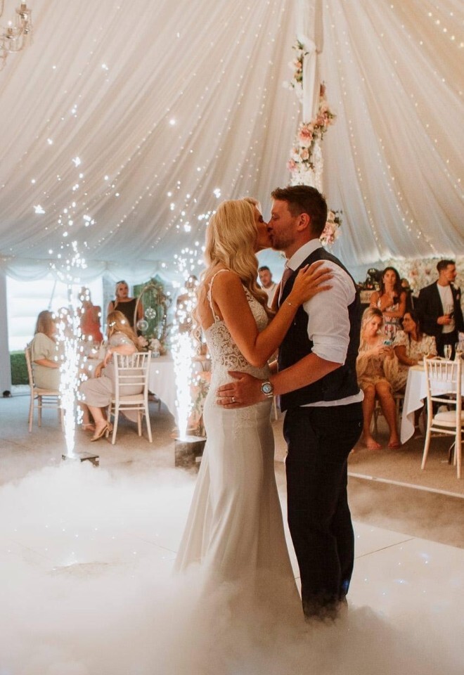 A newlywed couple kissing during their first dance on a dance floor covered in low-lying fog, surrounded by indoor sparkler fountains and seated wedding guests under a decorated white marquee tent.