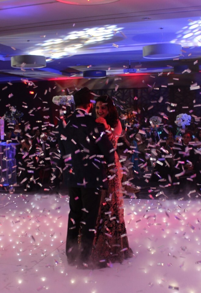 A couple sharing their first dance on a lit white dance floor covered in low fog, with silver confetti raining down around them under ambient venue lighting.