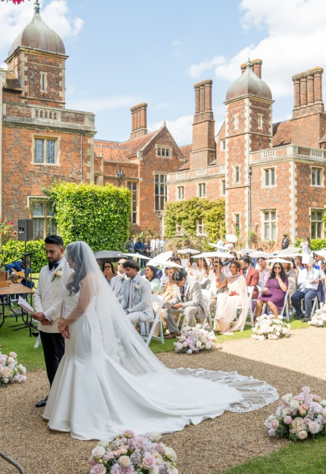 Outdoor wedding ceremony taking place on the grounds of North Mymms Park, showing a bride and groom at the altar framed by a stone arch pillar, with seated guests holding white umbrellas and the histo