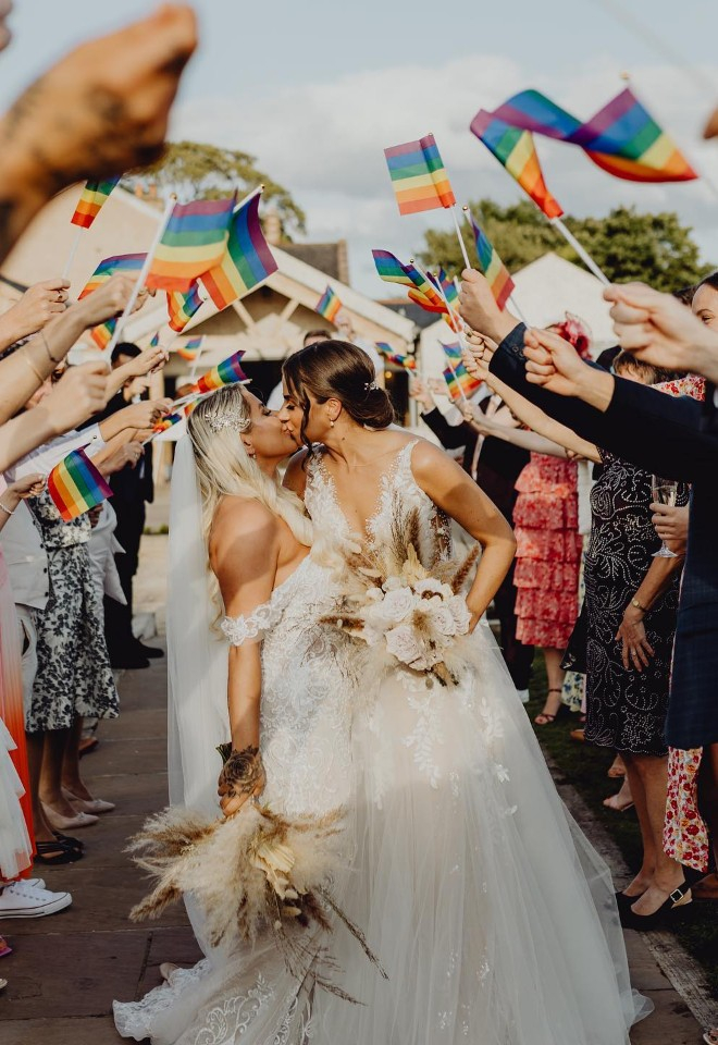 Same sex female wedding at Eden Wedding Barn, Cumbria kissing under an arch of lgbtq+ flags being held by their wedding guests