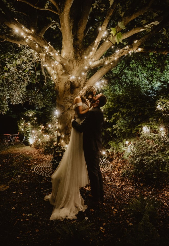 A couple sharing a romantic kiss under a tree decorated with fairy lights during their evening wedding, photographed by V & H Photography in Oxfordshire.