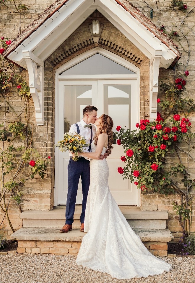 A bride and groom on the steps of the wedding venue covered in red roses, the bride in a lace gown holding a bouquet. Image by Sam Rundle Photography.