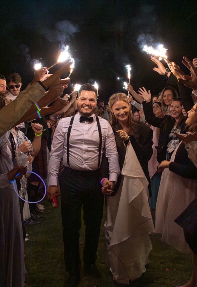 Bride and groom leaving the wedding party with family and friends forming a tunnel of sparklers in the evening